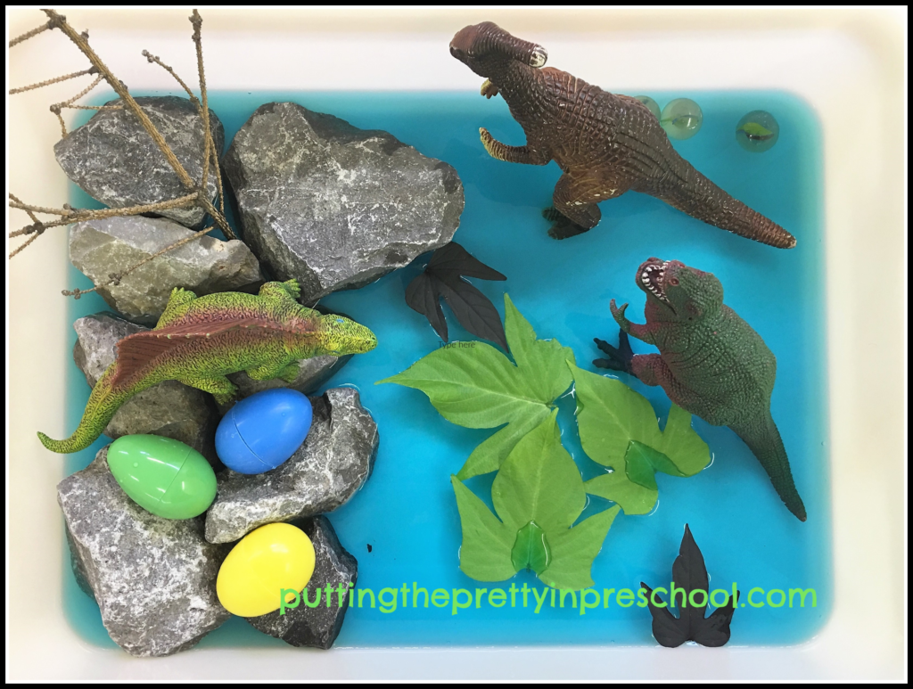 Small world scene with dinosaurs, rocks, twig, plastic eggs, marbles, blue water, and sweet potato vine leaves.