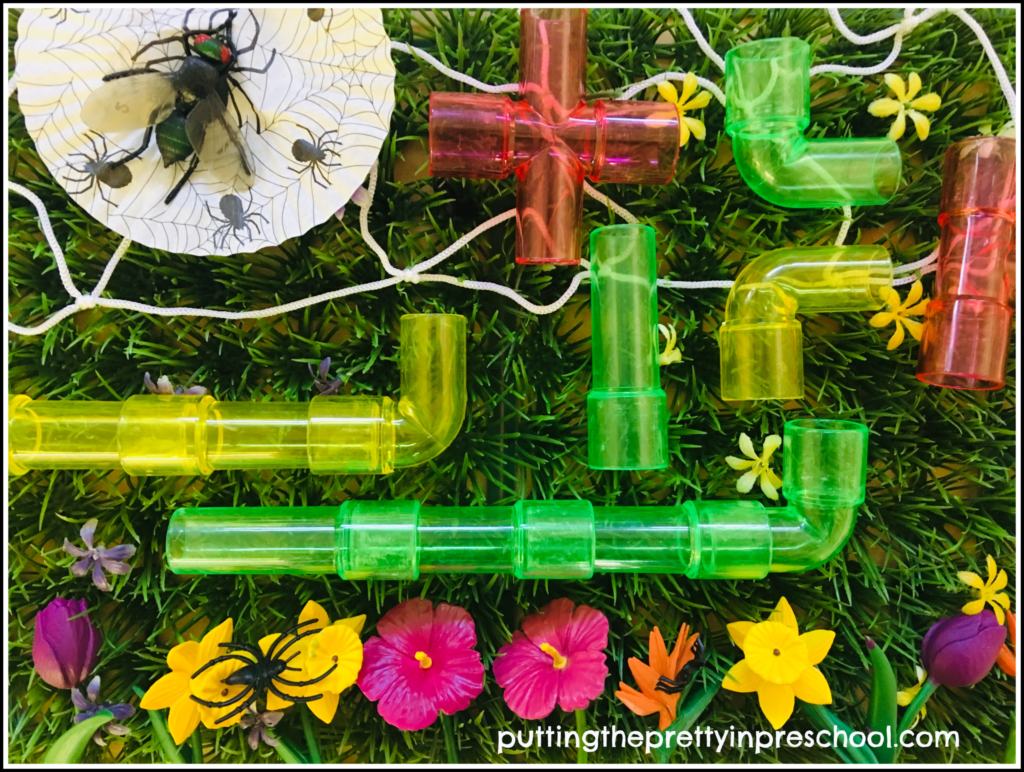 Spider themed small world with translucent pipe builders, spiders, a fly, flowers, and a spider web on a patch of grass.