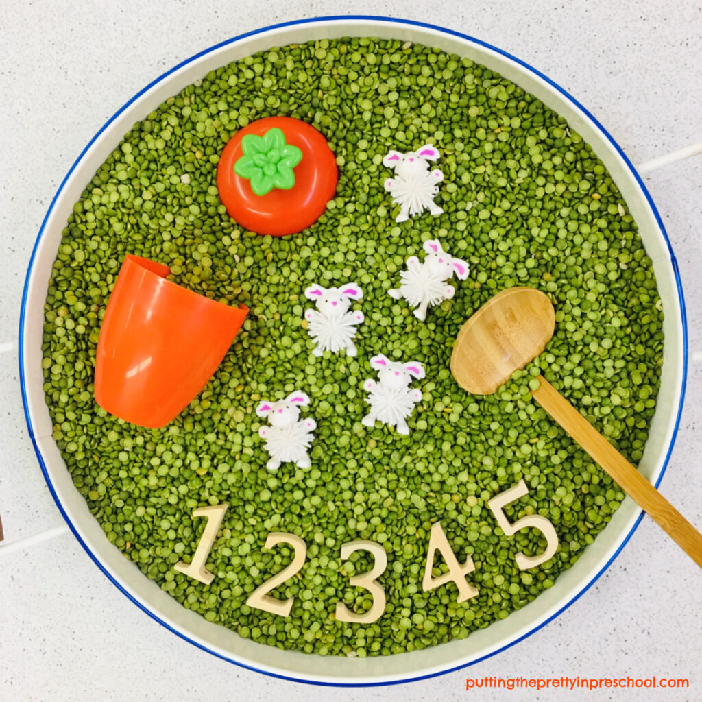 Split pea-based sensory tub with bunny woolies, numbers, ladle, and carrot cup with a lid.