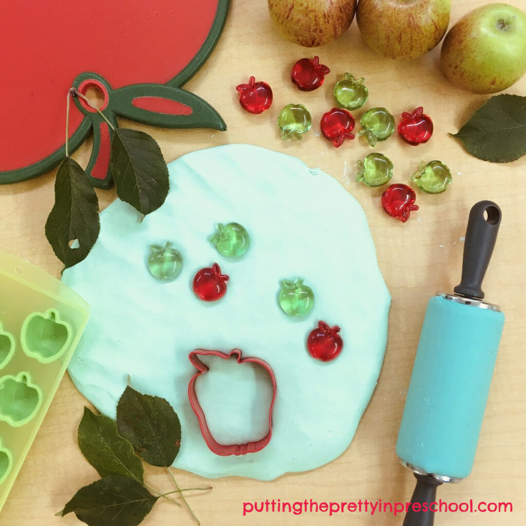 Apple themed playdough invitation with apple cutting board, ice cube tray, cookie cutters, leaves, and gems.