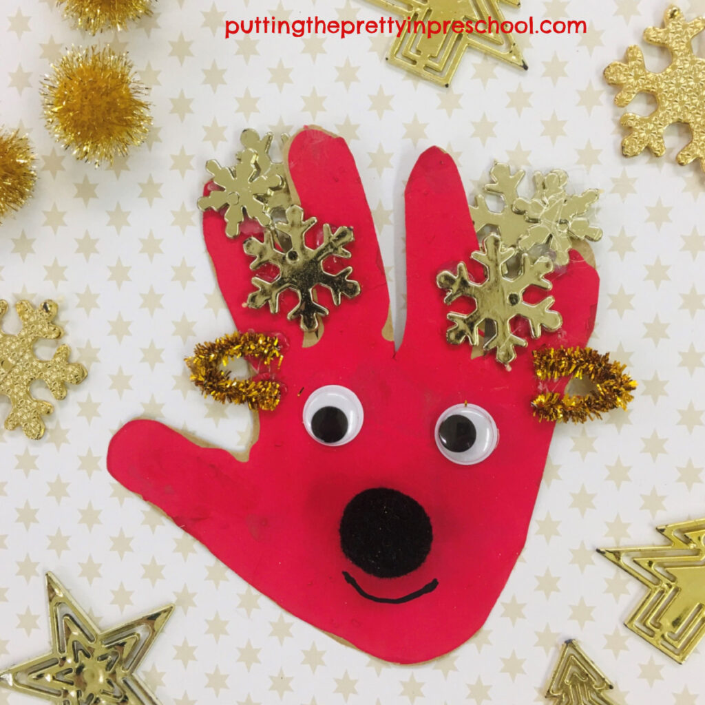 A red handprint turned into a friendly reindeer craft by adding snowflake antlers, wiggly eyes, pipe cleaner ears, and a pompom nose.
