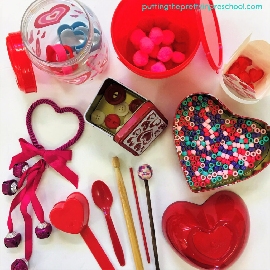 A collection of Valentine's Day-themed containers with loose parts and drumsticks for music-making.