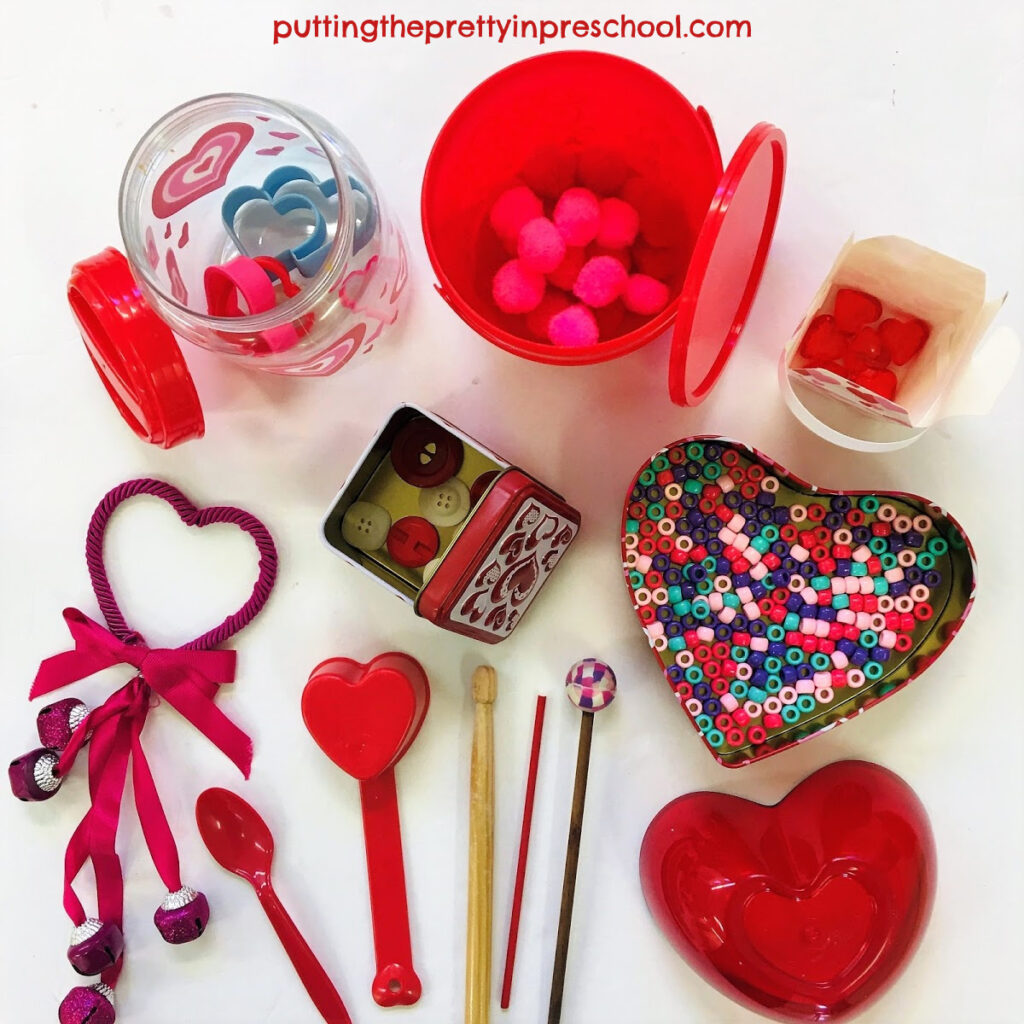 A collection of Valentine's Day-themed containers with loose parts and drumsticks for music-making.