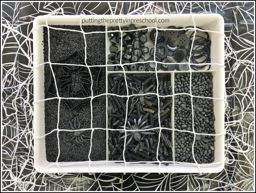 An all-black spider web sensory bin with noodles, rice, beans, spiders, and loose parts.
