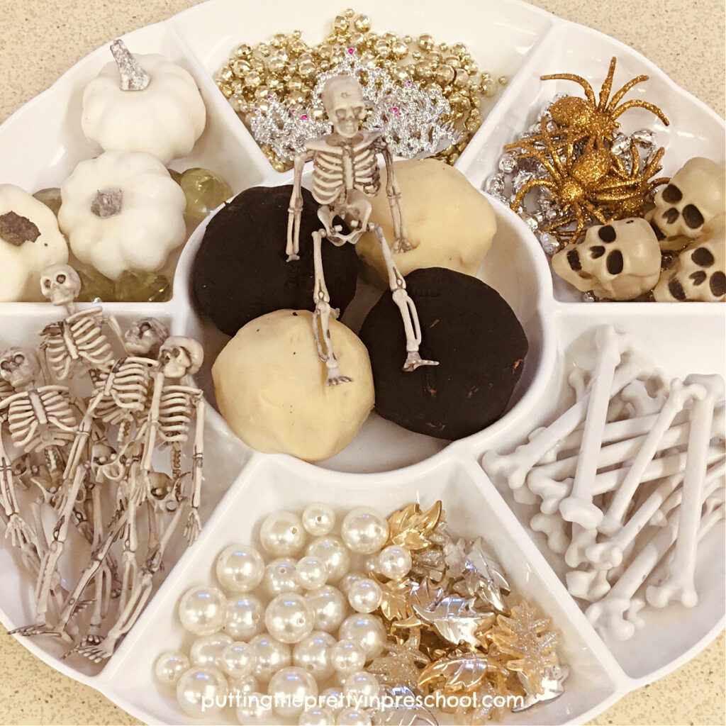 A skeleton sits atop black and white playdough in this inviting party tray. Other loose parts include pumpkins, spiders, skulls, and bones.