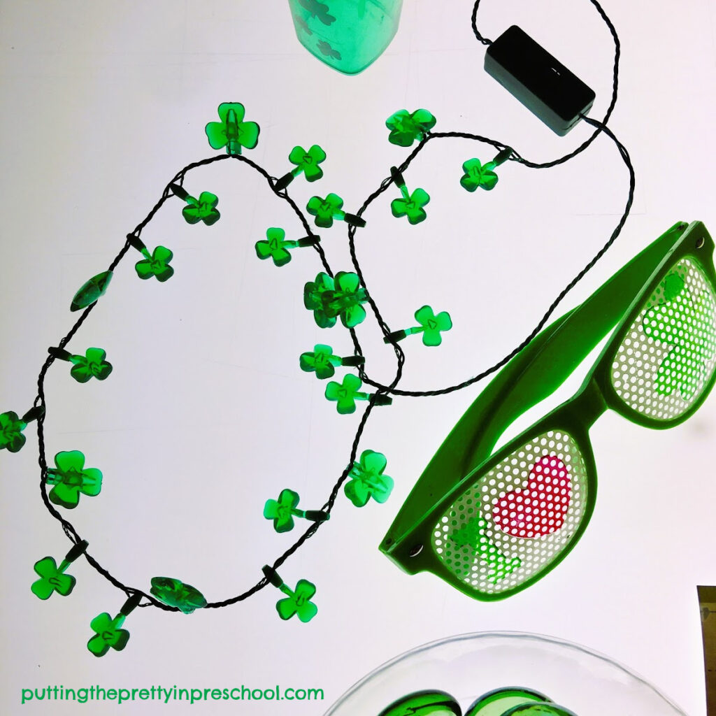 A green shamrock light-up necklace and pair of glasses are highlights in a St. Patrick's Day light table center.