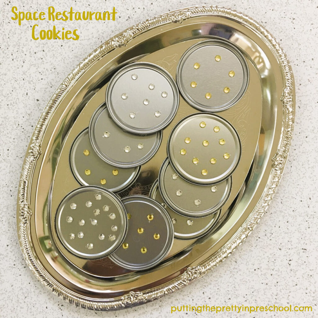 Round adhesive gem stickers added to juice lids make great chocolate chip cookies in a space restaurant pretend play center.