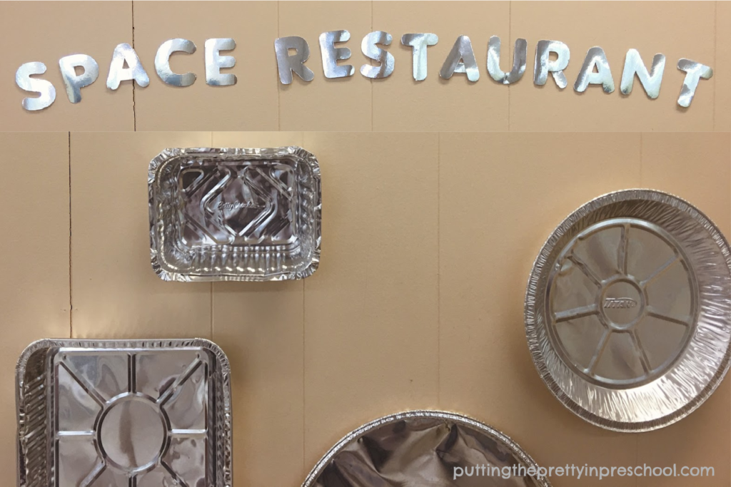 SIlver foil letters and aluminum foil containers and pans taped on a wall add ambiance to a space restaurant dramatic play center.