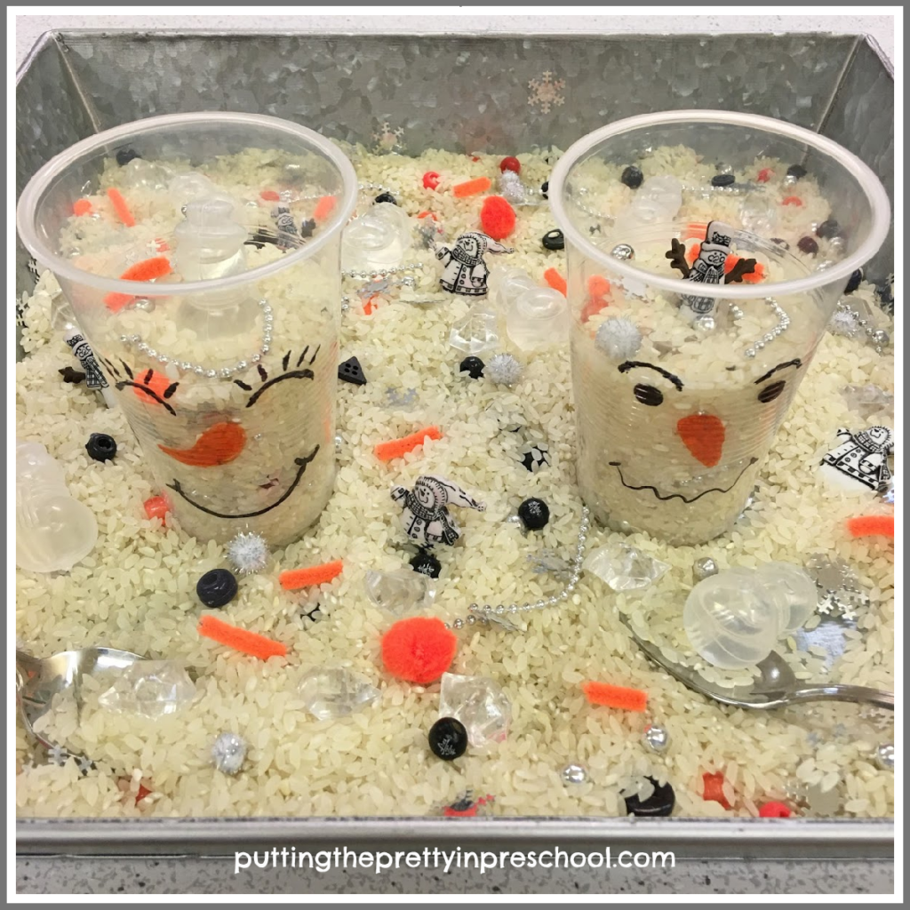 Snowman sensory tray with a rice base. Clear cup snowmen, themed buttons, and snowmen ice cubes are included along with clear, orange, and black bits and baubles.